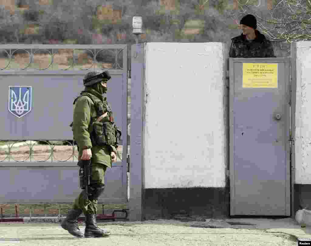 A Ukrainian serviceman stands guard at a Ukrainian military base as a uniformed man, believed to be a Russian serviceman, walks nearby in Perevalnoye outside Simferopol, March 6, 2014.