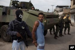 A masked police officer stands with a suspect in handcuffs, next to Brazilian marines during a surprise operation in Kelson's slum in Rio de Janeiro, Brazil, Feb. 20, 2018.